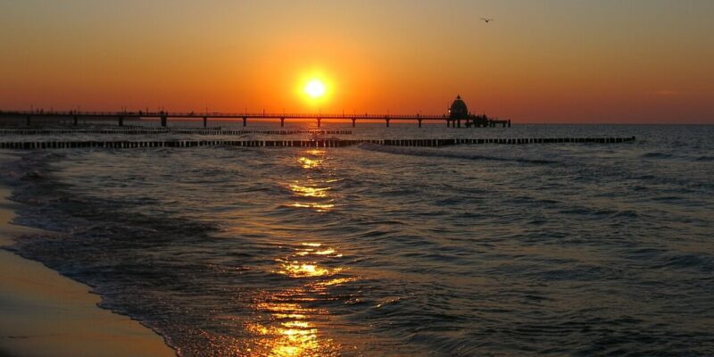 Ostseestrand Zingst Ein ruhiger Sonnenuntergang über dem Ostseestrand Zingst. Die tief am Himmel stehende Sonne wirft einen goldenen Widerschein auf das Wasser. Ein Pier ragt ins Meer und zeichnet sich als Silhouette gegen die warmen Farben des Himmels ab, während sanfte Wellen an die Küste plätschern.