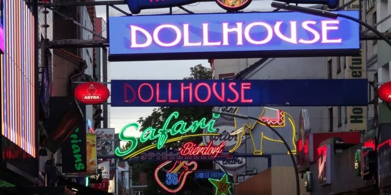 Reeperbahn Eine belebte Straße mit zahlreichen leuchtenden Neonschildern, darunter Dollhouse und Safari, erhellt die Nacht auf der Reeperbahn in Hamburg. Diese lebendige Szene fängt die farbenfrohe Atmosphäre eines urbanen Nachtlebenviertels voller Energie und Aufregung ein.