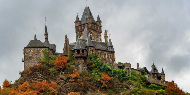 Die Reichsburg Cochem, eine mittelalterliche Burg mit zahlreichen Türmen und Spitzen, liegt auf einem felsigen Hügel. Die umliegenden Bäume leuchten in den Herbstfarben Orange, Rot und Gelb. Der Himmel ist bedeckt und wirft einen grauen Farbton über die Szenerie.