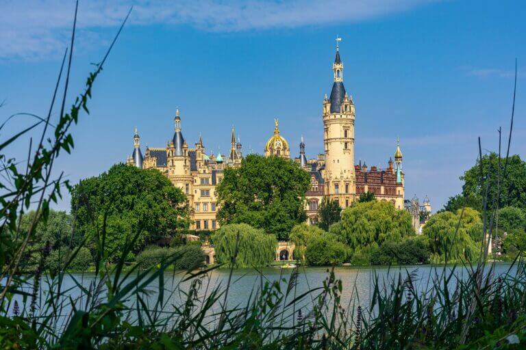Ein malerischer Blick auf Schloss Schwerin mit seinen verzierten Türmen und Spitzen vor einem klaren blauen Himmel. Im Vordergrund sind üppiges Grün und ein ruhiger See zu sehen, was das märchenhafte Aussehen des Schlosses noch verstärkt und es zu einem der bezauberndsten Schlösser in Mecklenburg-Vorpommern macht.