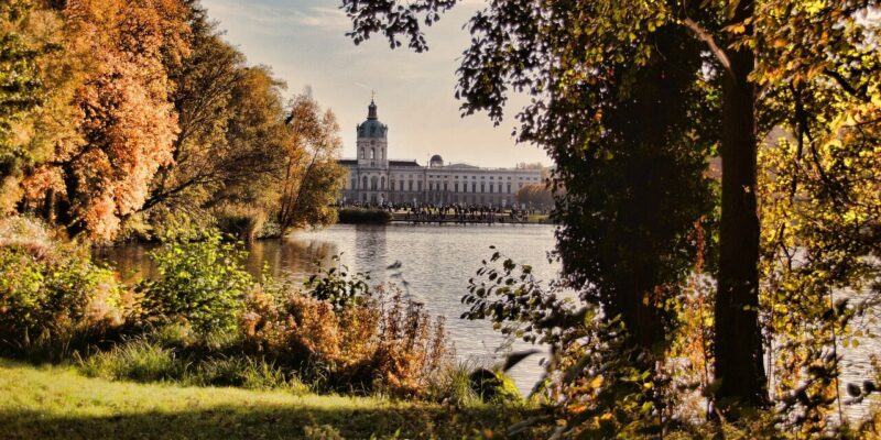 In der Ferne, jenseits eines ruhigen Sees, erhebt sich Schloss Charlottenburg als großes historisches Gebäude, eingerahmt von Bäumen mit herbstlichem Laub in Gold- und Orangetönen. Das Sonnenlicht fällt hindurch und schafft eine ruhige und malerische Szene.
