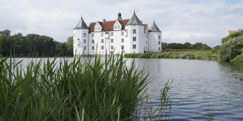 Schloss Glücksburg, ein weißes Schloss mit spitzen Türmen und rotem Dach, liegt an einem ruhigen See. Hohes grünes Schilf säumt den Vordergrund, während im Hintergrund unter einem wolkenblauen Himmel üppige Bäume zu sehen sind.
