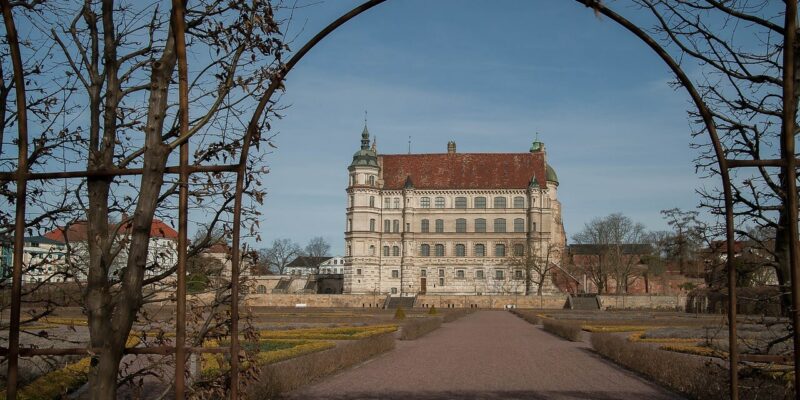 Schloss Güstrow ist ein prachtvolles historisches Gebäude mit einem roten Ziegeldach und wird von einem dekorativen Torbogen eingerahmt. Im Vordergrund führt ein von gepflegten Gärten gesäumter Weg zum Eingang. Der Himmel ist klar und blau und unterstreicht die zeitlose Eleganz dieses architektonischen Wunders.