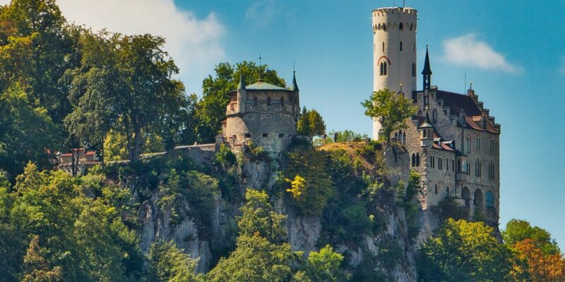 Schloss Lichtenstein thront auf einem felsigen Hügel und ist von üppigem Grün unter einem klaren blauen Himmel umgeben. Das Schloss verfügt über einen hohen, runden Turm und mehrere kleinere Gebäude mit steilen Dächern. Bäume rahmen die Szene ein und verstärken die malerische Umgebung.