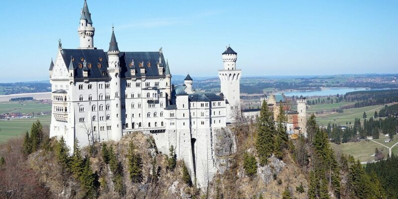 Ein malerischer Blick auf Schloss Neuschwanstein, das auf einer von Bäumen umgebenen Klippe thront. Das Schloss zeichnet sich durch hohe Türme und eine helle Fassade aus, vor einer Kulisse aus Feldern, Wäldern und einem entfernten See unter einem klaren blauen Himmel.
