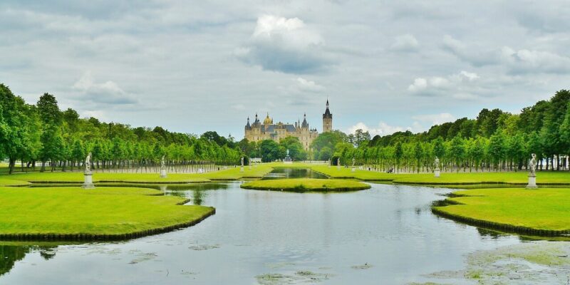 Eine malerische Aussicht auf Schloss Schwerin, ein prachtvolles Schloss mit mehreren Türmen in der Ferne. Es ist von üppigen grünen Gärten umgeben und hat im Vordergrund einen großen, reflektierenden Teich. Der Himmel ist mit Wolken bedeckt, was zu seiner majestätischen Anziehungskraft beiträgt.