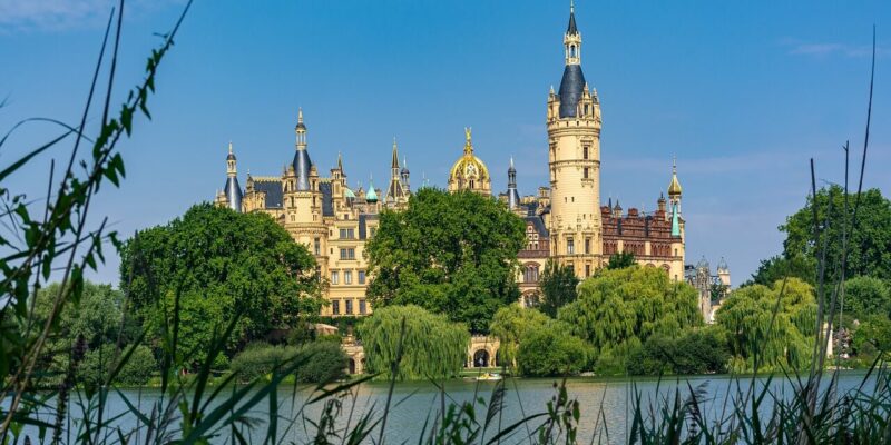 Ein prachtvolles, reich verziertes Schloss mit mehreren Türmen und einem hohen Turm in der Mitte, das an das Schweriner Schloss erinnert, steht umgeben von üppigem Grün. Das majestätische Bauwerk spiegelt sich in einem ruhigen See im Vordergrund unter einem klaren blauen Himmel.