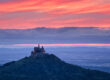 Ein malerisches Schloss, das an die Burg Hohenzollern erinnert, thront auf einem bewaldeten Hügel und hebt sich vor einem dramatischen Sonnenuntergangshimmel mit leuchtenden Rosa- und Orangetönen ab. Im Hintergrund erstrecken sich sanfte Hügel und ferne Landschaften und schaffen eine ruhige und majestätische Szene, die typisch für Sehenswürdigkeiten in Baden-Württemberg ist.