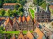 Luftaufnahme von Lübeck, einem Highlight unter den Sehenswürdigkeiten in Schleswig-Holstein, mit mittelalterlicher Architektur mit roten Backsteingebäuden und dem ikonischen Holstentor. Die alten Lagerhäuser und grünen Rasenflächen sind von Straßen und Bäumen unter einem klaren Himmel umgeben.