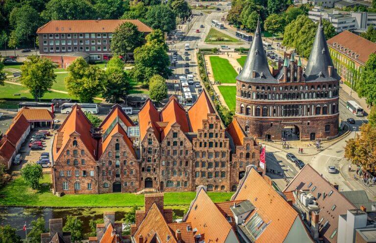 Luftaufnahme von Lübeck, einem Highlight unter den Sehenswürdigkeiten in Schleswig-Holstein, mit mittelalterlicher Architektur mit roten Backsteingebäuden und dem ikonischen Holstentor. Die alten Lagerhäuser und grünen Rasenflächen sind von Straßen und Bäumen unter einem klaren Himmel umgeben.