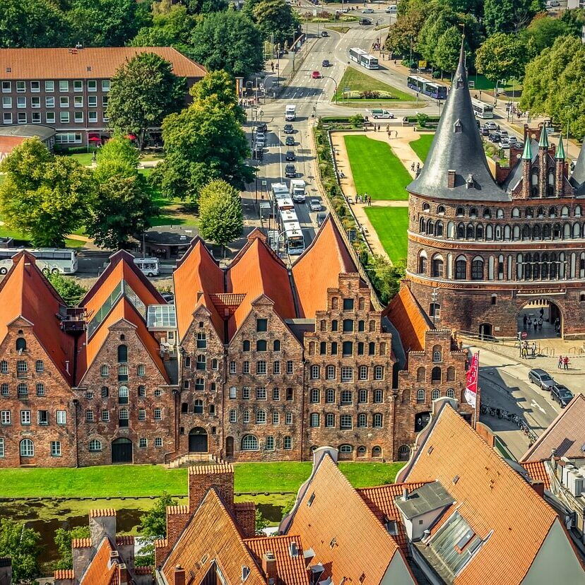 Luftaufnahme von Lübeck, einem Highlight unter den Sehenswürdigkeiten in Schleswig-Holstein, mit mittelalterlicher Architektur mit roten Backsteingebäuden und dem ikonischen Holstentor. Die alten Lagerhäuser und grünen Rasenflächen sind von Straßen und Bäumen unter einem klaren Himmel umgeben.