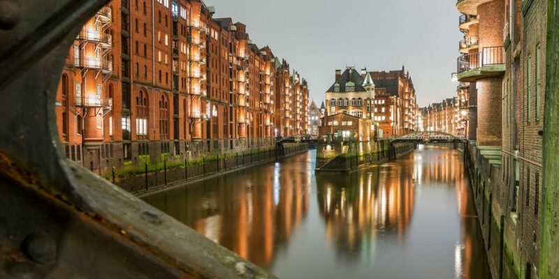 Speicherstadt Eine nächtliche Szene der Speicherstadt, einem historischen Viertel mit beleuchteten roten Backsteingebäuden, die einen ruhigen Kanal säumen und das warme Licht reflektieren. In der Mitte des Kanals steht ein Gebäude, das von einer Metallbrücke aus sichtbar ist.