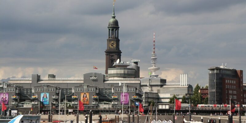 St. Michaelis Kirche Eine Stadtansicht mit einer Uferpromenade und angedockten Booten. Im Hintergrund erhebt sich die St. Michaelis Kirche in Hamburg über modernen Gebäuden. Flaggen schmücken die Gegend und der Himmel ist mit Wolken bedeckt.