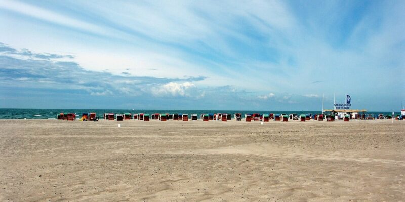 Strand von Warnemünde Am Strand von Warnemünde liegt ein breiter Sandstrand mit verstreuten Strandkörben am Meer. Der Himmel ist teilweise bewölkt und weist blaue und weiße Streifen auf. In der Ferne, wo das Meer unter einem weiten Himmel auf den Horizont trifft, erwartet Sie Ruhe.
