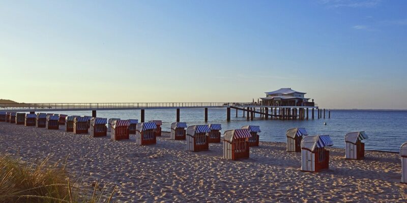 Am Timmendorfer Strand an der Ostsee entfaltet sich bei Sonnenuntergang eine ruhige Strandszene mit Reihen gestreifter Strandstühle im Sand. Ein langer Pier erstreckt sich über ruhiges blaues Wasser und führt zu einem charmanten Gebäude auf Stelzen. Der Himmel ist klar und in sanften Blau- und Rosatönen gemalt.