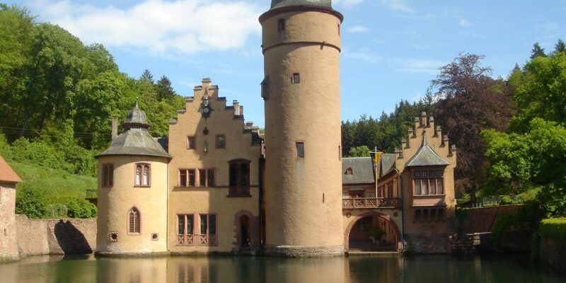 Schloss Mespelbrunn: Wasserschloss mit hohem Rundturm und Treppengiebeln, das sich in der ruhigen Wasseroberfläche des umgebenden Weihers spiegelt, eingebettet in bewaldete Hügel des Spessarts.