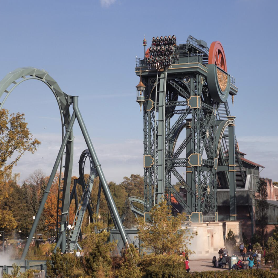Ein Achterbahnzug steht kurz vor dem vertikalen Absturz auf der Spitze eines historischen Minenturms, daneben ist ein großer Looping zu sehen.