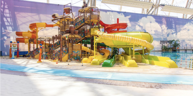 Jungle Splash Wasserspielplatz für Kinder Der farbenfrohe Kinder-Wasserspielplatz Jungle Splash im Tropical Islands mit mehreren kleinen Rutschen, Kletterelementen und Wassereimern vor einer künstlichen Wolkenwand.