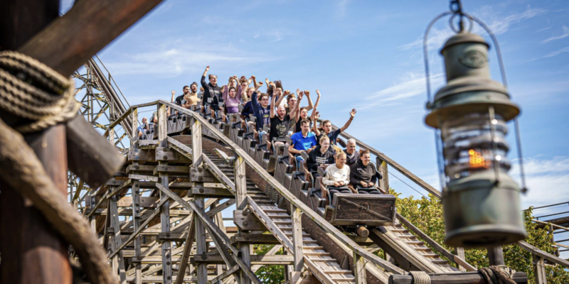 Eine vollbesetzte Holzachterbahn der Attraktion Colossos im Heide Park fährt bei strahlendem Sonnenschein eine Schiene hinab. Die Fahrgäste haben jubelnd die Arme in die Luft gestreckt. Im Vordergrund ist unscharf eine rustikale Holzlaterne zu sehen.