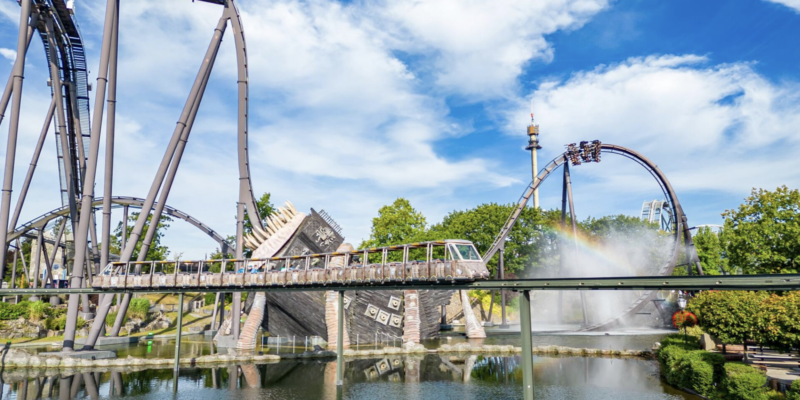 Panoramaansicht im Heide Park mit der Achterbahn Krake und dem Monorail. Im Vordergrund fährt die Einschienenbahn über einen See, dahinter ragt das Wrack eines Piratenschiffs aus dem Wasser. Im Hintergrund sind die Schienen des Dive Coasters Krake sowie eine Wasserfontäne mit einem kleinen Regenbogen zu sehen.
