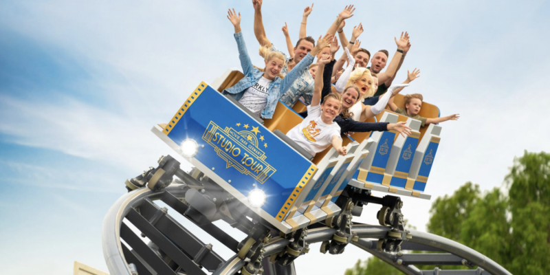 Eine Gruppe fröhlicher Menschen sitzt in den Wagen der Achterbahn "Movie Park Studio Tour" und streckt während der Fahrt die Arme begeistert in die Luft.