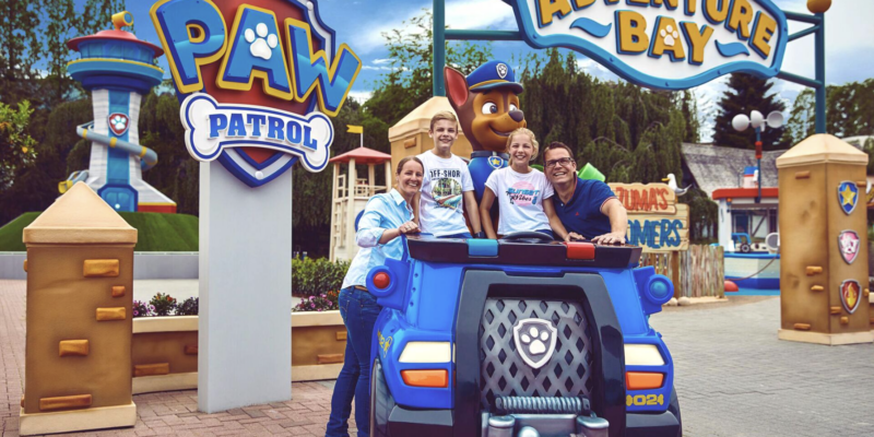 Eine Familie posiert lächelnd für ein Foto im "Adventure Bay"-Bereich des Movie Parks hinter einem blauen PAW-Patrol-Polizeiauto und neben einer großen Chase-Figur.